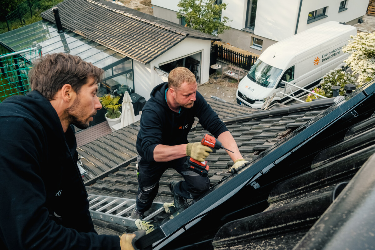 Zwei Menschen auf einem Dach bringen eine Solaranlage an. Im Hintergrund ein Sprinter von Energiekonzepte Deutschland.