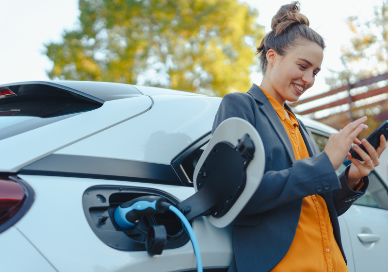 Frau, die vor einem elektrischen Auto mit Handy in der Hand steht