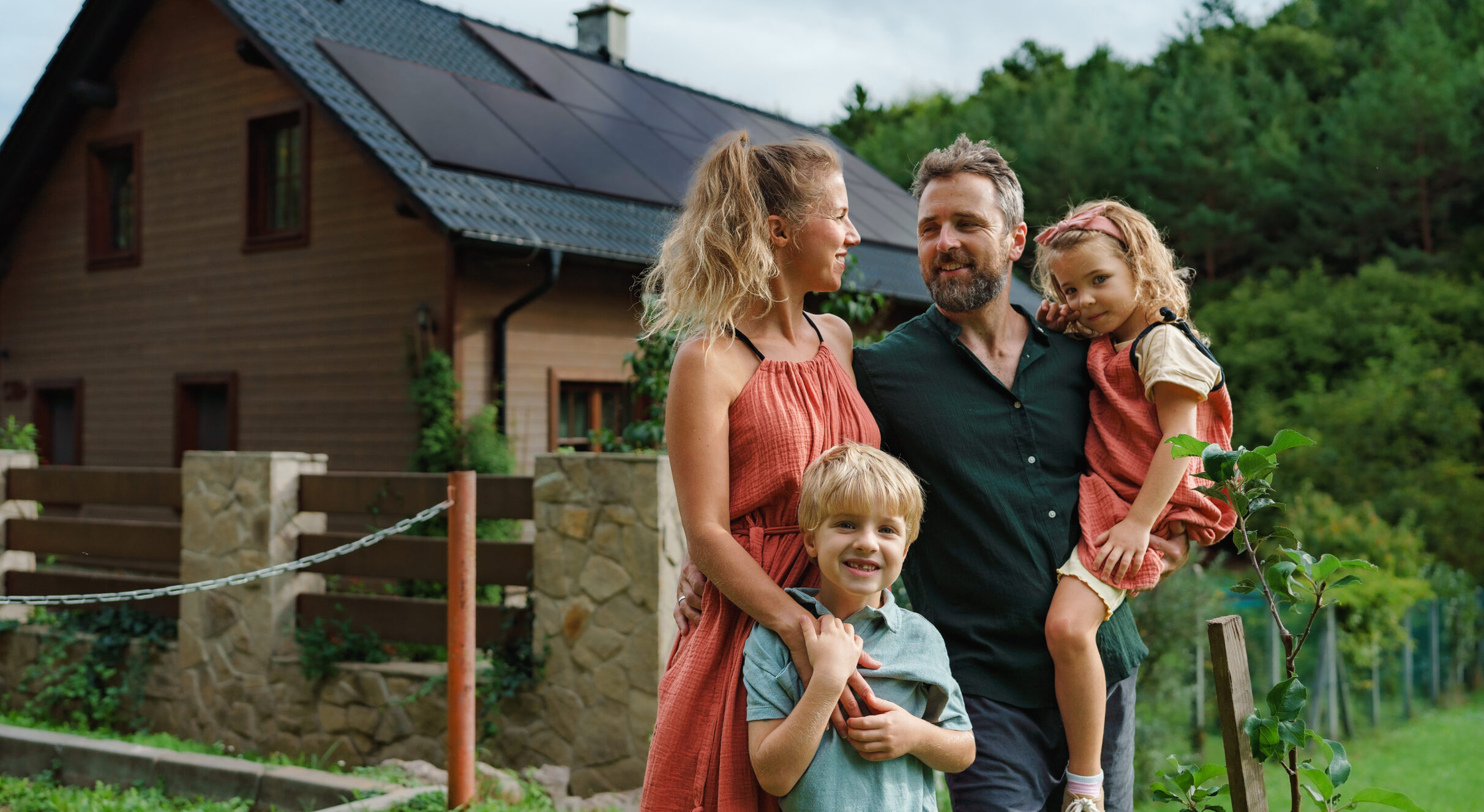 Familie aus Vater, Mutter, Tochter & Sohn vor Einfamilienhaus mit Solaranlage auf dem Dach.